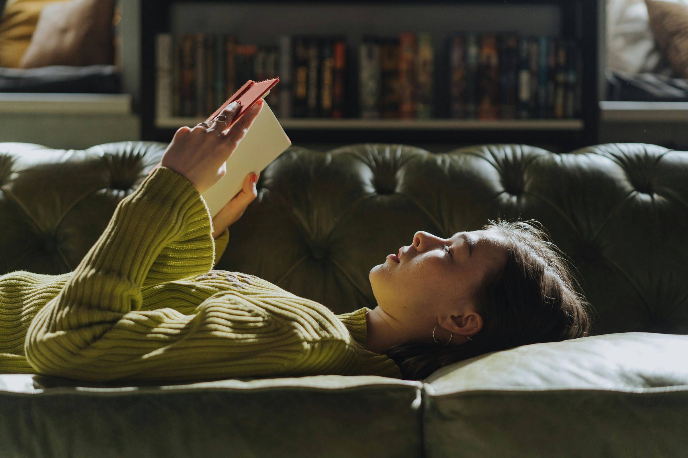 Young woman in a lime green sweater reading a book on a dark green tufted sofa.