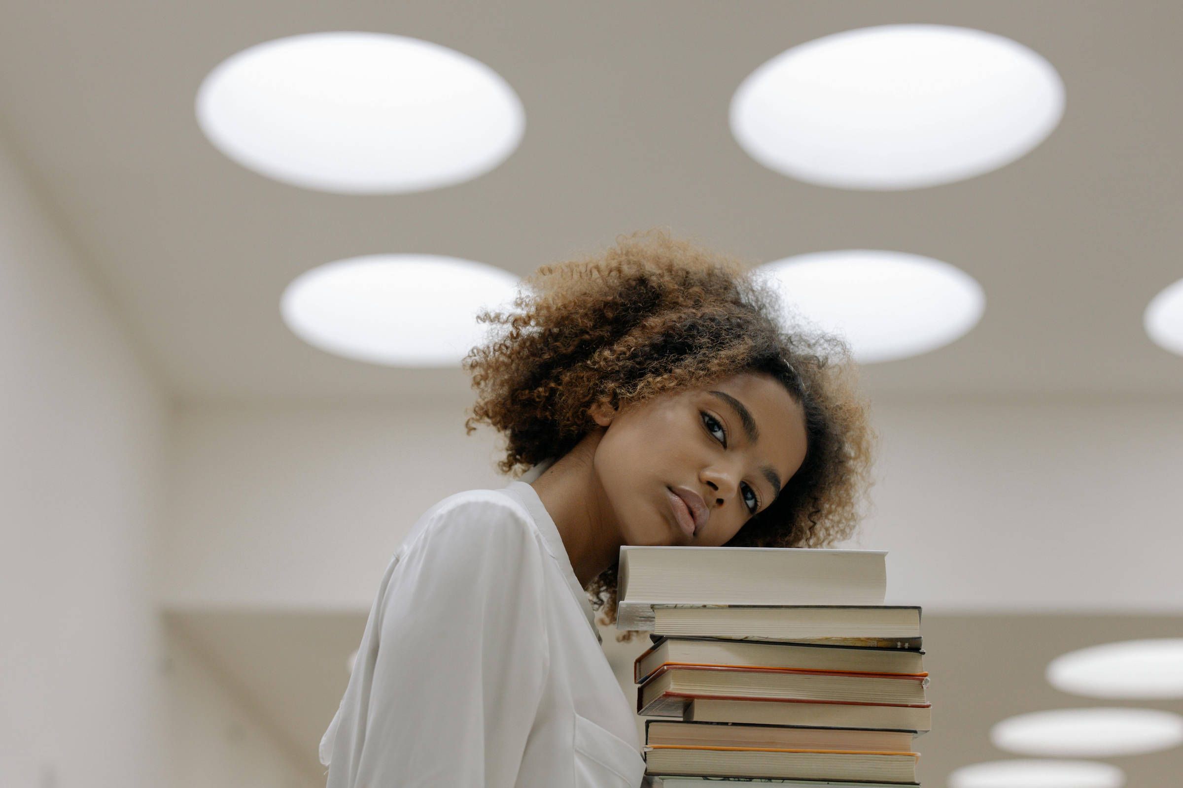 A woman in white leans her head on a stack of books, with round ceiling lights in the background.