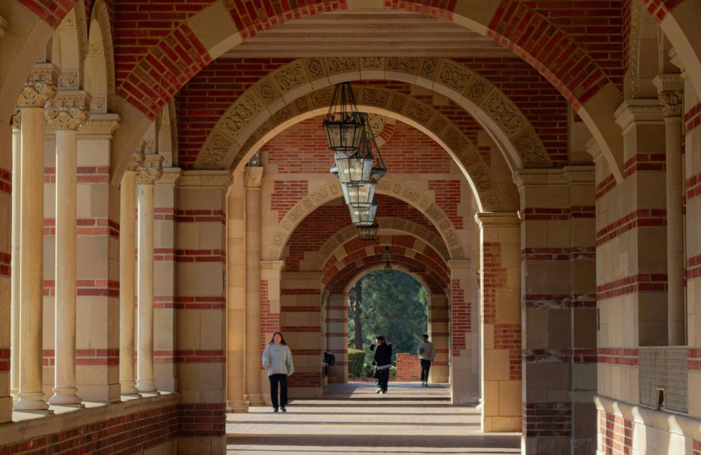 Brick archway corridor with hanging lanterns and sunlight casting shadows, people walking in the distance.