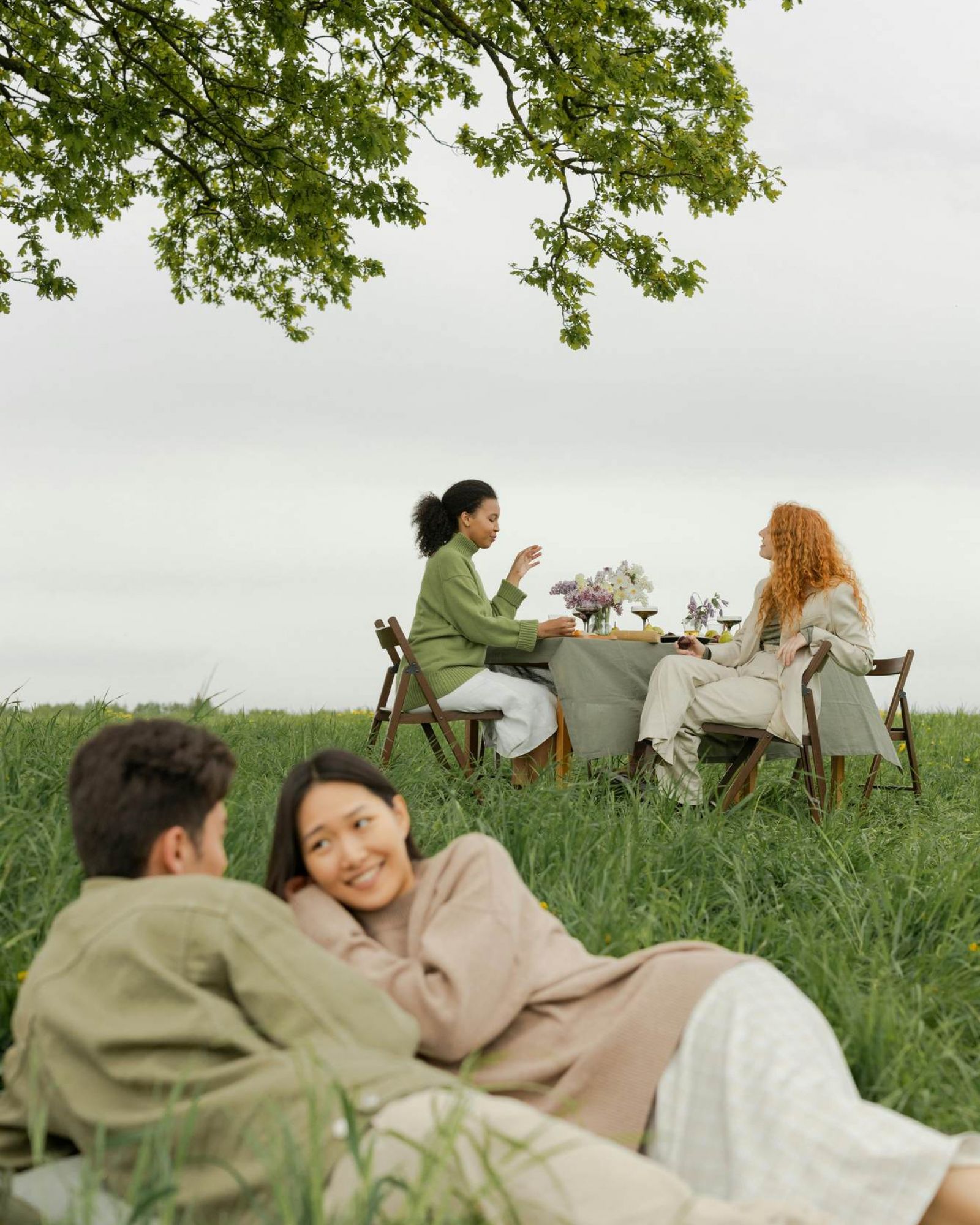 Two people lie in tall grass while two others sit at a table outdoors, talking under a tree.