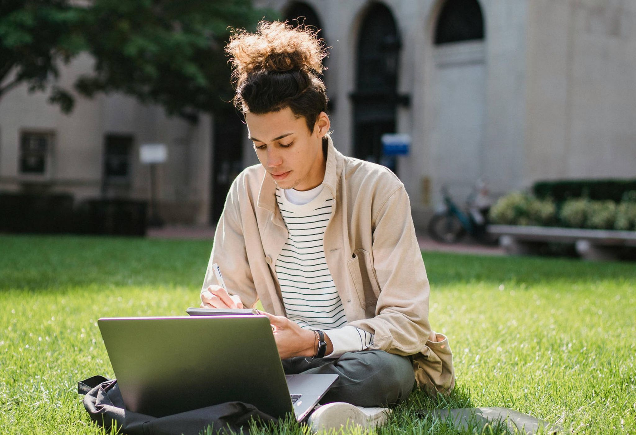 Person sitting on grass with a laptop, writing in a notebook, with a building in the background.