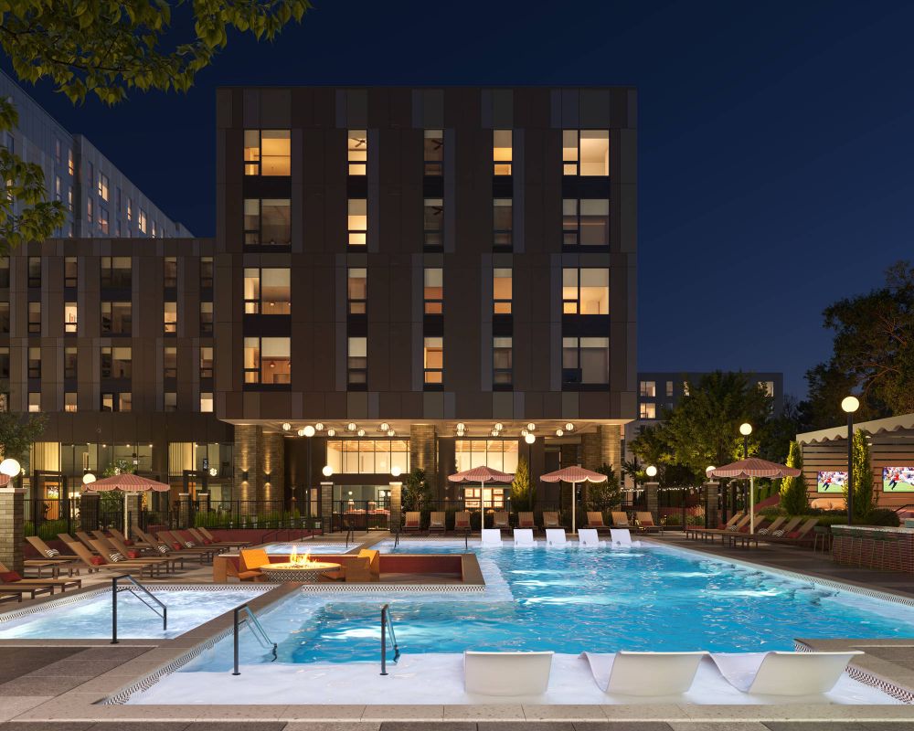 Night view of a modern apartment building with a lit outdoor pool, lounge chairs, and a firepit seating area.