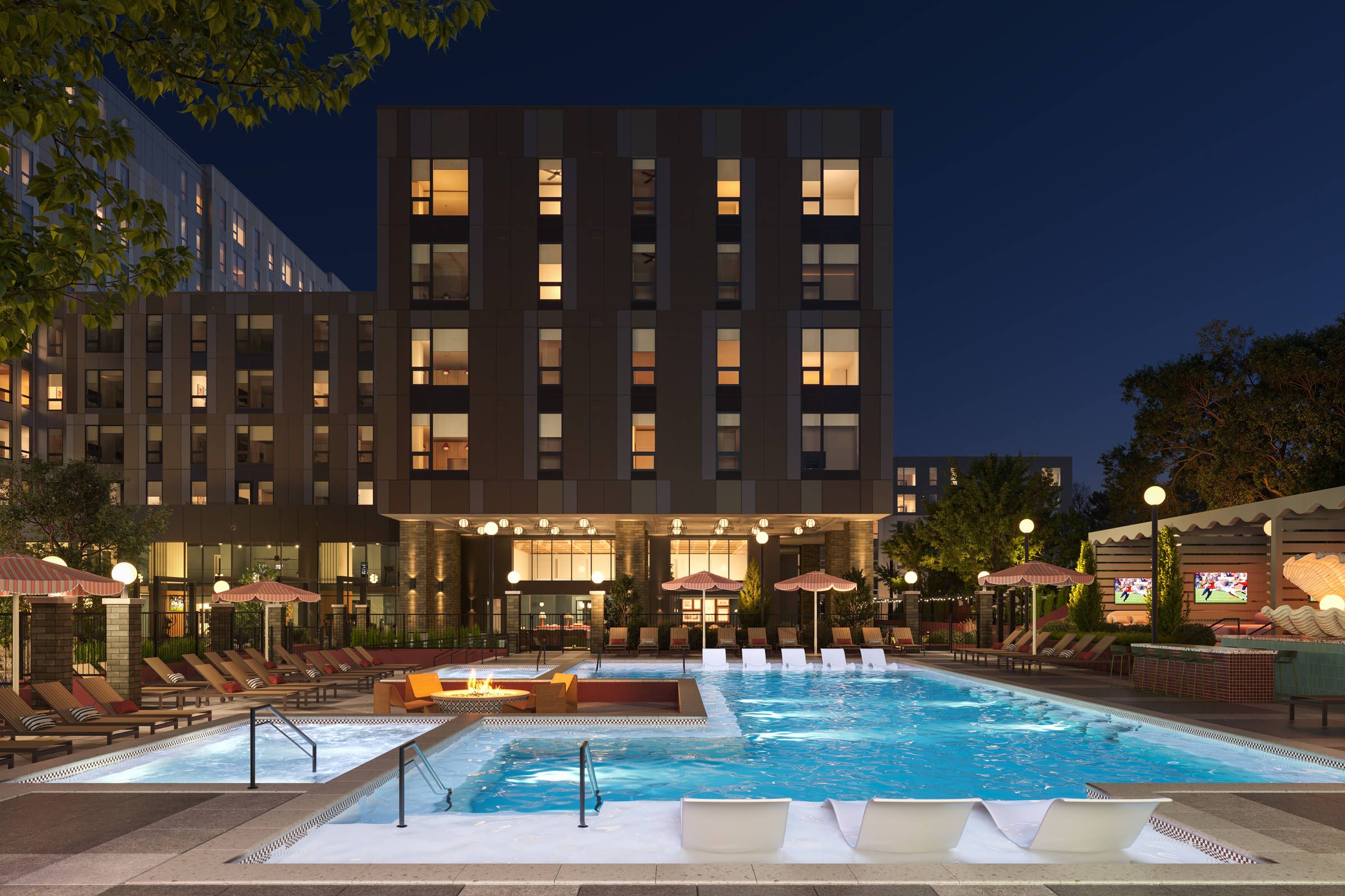 Night view of a modern apartment building with a lit outdoor pool, lounge chairs, and a firepit seating area.