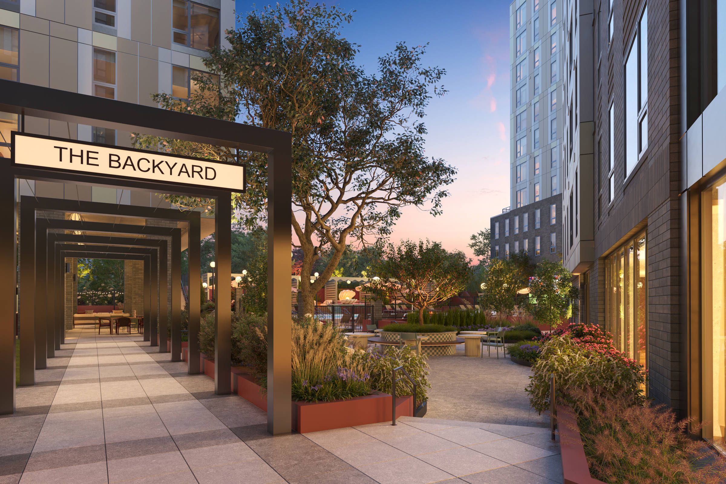 Modern apartment courtyard at sunset with "The Backyard" sign, greenery, seating, and decorative lighting.