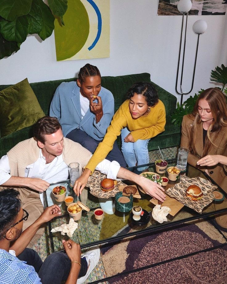 Five friends sit around a coffee table, sharing food and drinks in a cozy, modern living room.